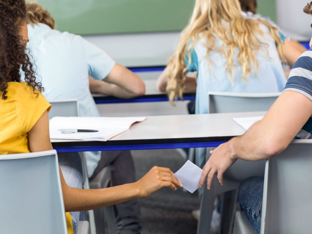 Une élève avec un chandail jaune glisse subtilement une note dans la main de son camarade de classe.