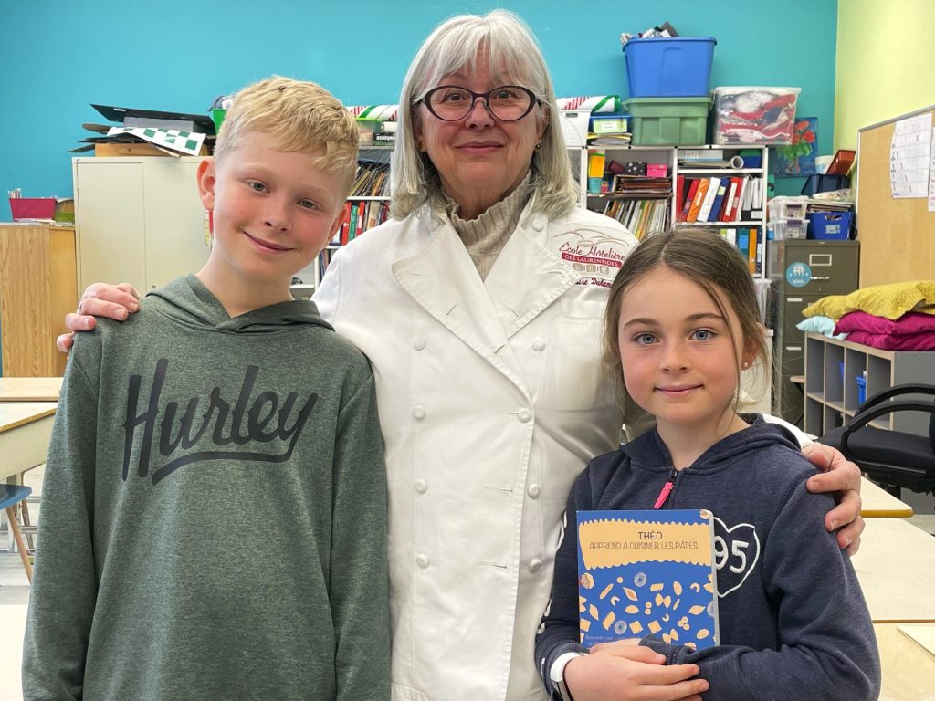 Louise Duhamel est entourée de deux élèves de 4e année qui présentent son livre Théo apprend à cuisiner les pâtes